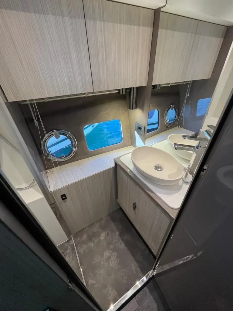 Draufsicht auf das Badezimmer einer Cranchi T36 Yacht, die glänzende weiße Arbeitsplatten mit zwei Waschbecken und rechteckigen Spiegeln zeigt, beleuchtet durch natürliches Licht aus ovalen Bullaugen.