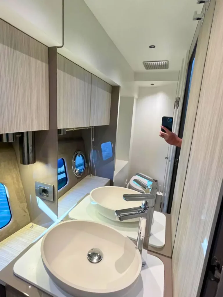 Innenraum eines Cranchi T36 Yachtbadezimmers mit holzverkleideten Schränken, zwei ovalen Bullaugen und einem weißen Waschbecken unter einem Spiegel.