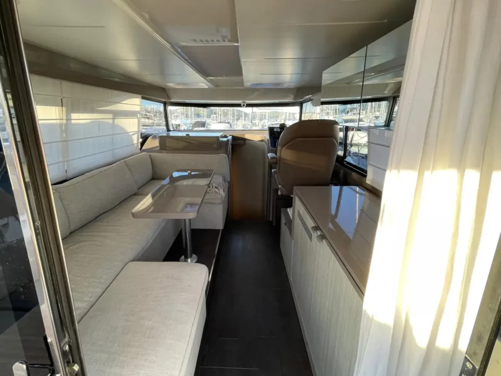 Geräumiger Innenraum einer Cranchi T36 Yacht mit beigefarbenen gepolsterten Sitzgelegenheiten und hellen Holzoberflächen, beleuchtet durch natürliches Licht aus großen Fenstern.