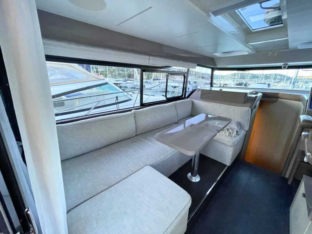 Innenansicht des Loungebereichs einer Cranchi T36 Yacht mit einem langen beigen Sofa, einem glänzenden Tisch und Panoramafenstern, die einen Blick auf den Yachthafen bieten.