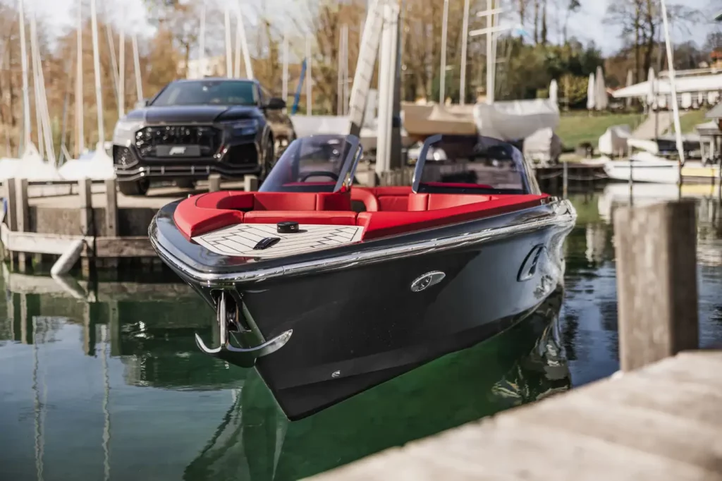 ABT Marian M 800-R Motorboot mit glänzend schwarzer Außenhaut und leuchtend rotem Interieur, vertäut an einem Steg neben Luxusautos und Segelbooten.