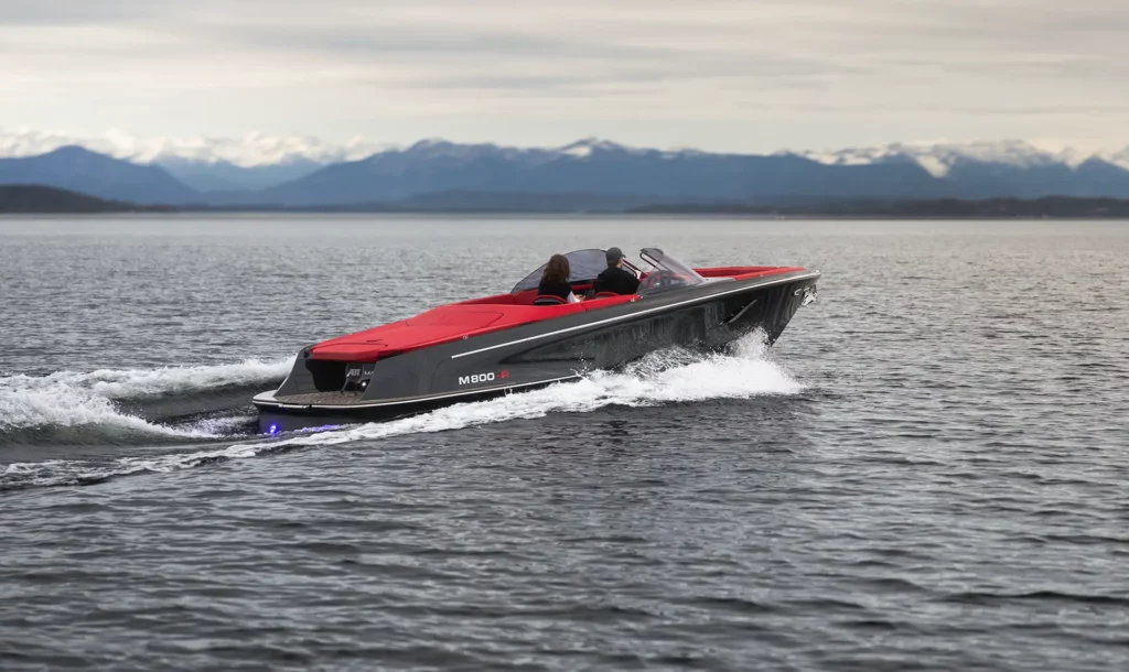 Ein Marian M 800-R mit rotem Deck und schwarzem Rumpf fährt auf einem ruhigen See vor einer malerischen Bergkulisse. Der Fahrer und ein Passagier sind sichtbar, während das Boot auf der Wasseroberfläche gleitet und leuchtende, blaue Lichter an seinem Heck hinterlässt.