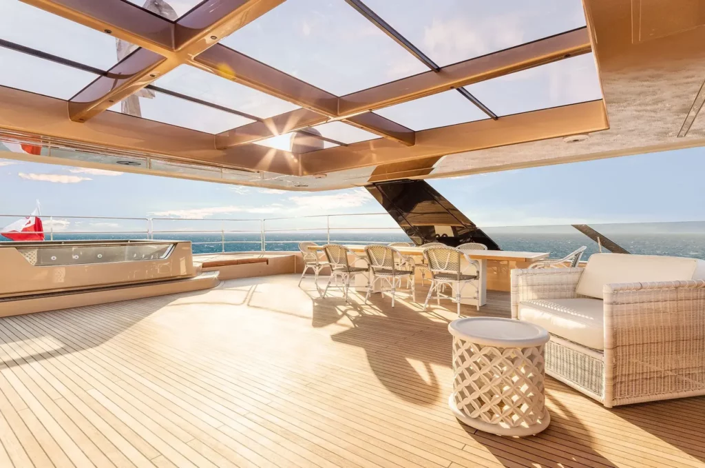 The top deck of the 100 Sunreef Power yacht, showcasing a sunlit seating area with white wicker furniture, a hot tub, and a bar area, with the ocean stretching out in the background.