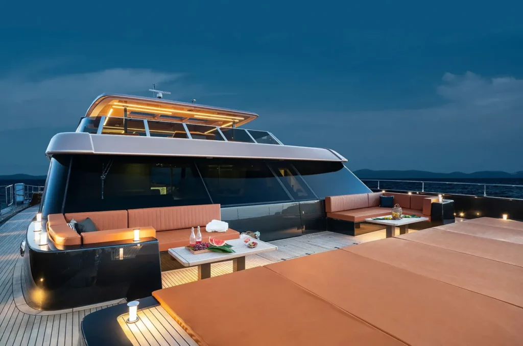 The upper deck of a 100 Sunreef Power luxury yacht at night, featuring ambient lighting, cushioned seating areas with tables, and panoramic ocean views.