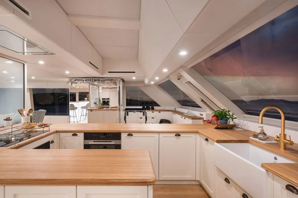 A modern, U-shaped kitchen on the 100 Sunreef Power yacht, featuring sleek wooden countertops, white cabinetry, and a gold faucet, with large windows offering a view of the ocean at sunset. The kitchen is well-lit with recessed lighting, and stylish bar stools can be seen in the background.