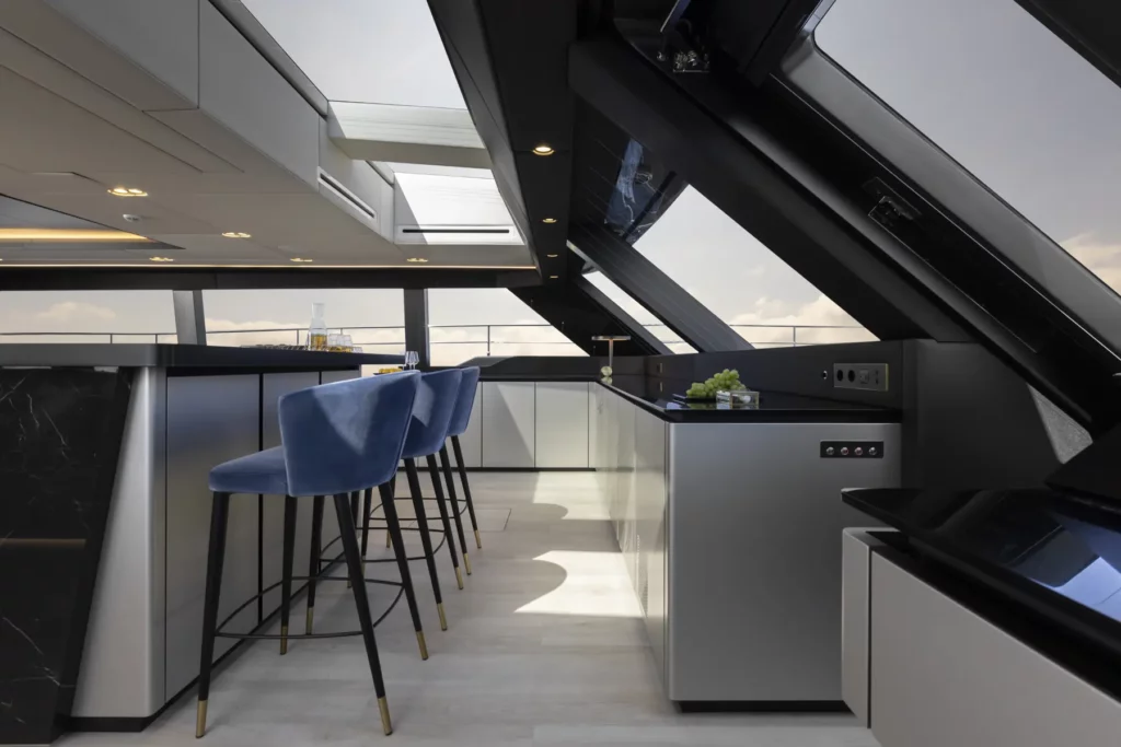 Ein moderner Barbereich im Inneren der 80 Sunreef Power Yacht, mit eleganten schwarzen Arbeitsplatten, blauen hohen Hockern und einem minimalistischen Design, das sich harmonisch in die umgebenden Sitzbereiche einfügt, alle mit großen Fenstern mit Blick auf das Meer.